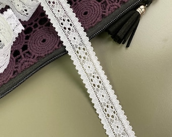 May include: White lace trim with a delicate floral pattern. The trim is shown on top of a purple crocheted bag.