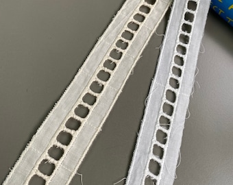 May include: Two pieces of white cotton lace trim with a square-shaped openwork design. The lace trim is lying on a gray surface.