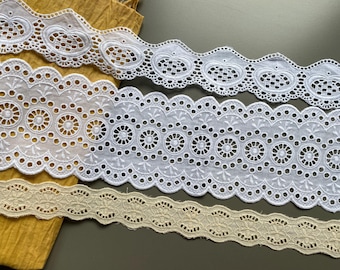 May include: Three white cotton lace trims with scalloped edges and different patterns. The top trim has a repeating oval pattern, the middle trim has a repeating circle pattern, and the bottom trim has a repeating floral pattern. A fourth trim is beige with a repeating floral pattern.