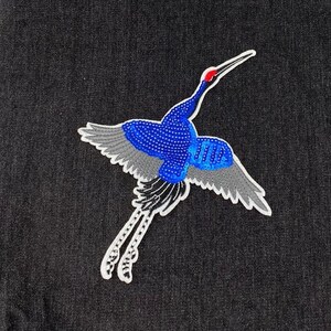 May include: A black denim tote bag with a blue and gray embroidered crane design. The bag has yellow, red, and blue patterned straps.
