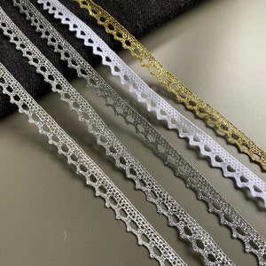 May include: Close-up of several decorative lace ribbons in silver, white, and gold. The ribbons have a scalloped edge and a repeating geometric pattern. The ribbons are arranged diagonally against a dark fabric and a grey surface.