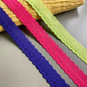 Puede incluir: Primer plano de tres cintas caladas de colores en azul, rosa intenso y verde lima. Cada cinta tiene un borde festoneado y un patrón floral repetitivo. Un tejido amarillo está en el fondo.