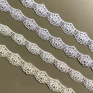 May include: Four rows of white lace trim with scalloped edges and floral designs. The lace is arranged in a staggered pattern on a brown background.