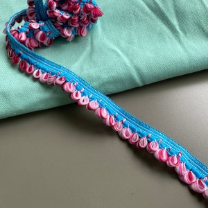 Könnte beinhalten: Nahaufnahme einer türkisfarbenen und rosafarbenen Borte mit einem gekräuselten Rand. Die Borte ist aus Stoff und hat einen zarten, femininen Look.