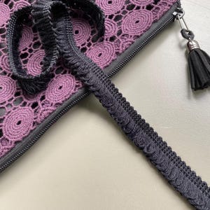 Könnte beinhalten: Nahaufnahme einer schwarzen und lila gehäkelten Reißverschlusstasche mit einem schwarzen Quasten-Reißverschlussanhänger. Eine schwarze dekorative Zierleiste mit einer geschlungenen Kante ist auf der Oberfläche platziert. Die Tasche hat ein kreisförmiges Muster.