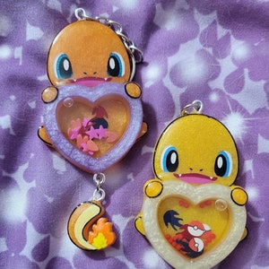 Könnte beinhalten: Zwei Schlüsselanhänger aus Harz mit Pokémon-Charakteren. Der erste Schlüsselanhänger ist ein hellorangefarbener Glurak mit einem lilafarbenen herzförmigen Anhänger, der mit Glitzer und rosa Sternen gefüllt ist. Der zweite Schlüsselanhänger ist ein gelber Pikachu mit einem weißen herzförmigen Anhänger, der mit Glitzer und roten Sternen gefüllt ist.