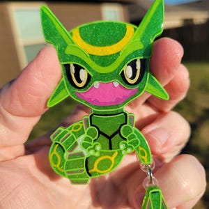 May include: A green and yellow glitter charm of a Pokemon character,  Sceptile, with a pink mouth and a clear chain attached to a smaller green glitter charm.