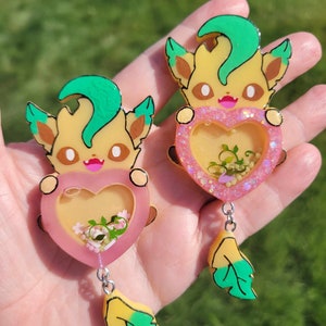 Puede incluir: Dos amuletos de personajes de dibujos animados amarillos y verdes con corazones rosas llenos de purpurina y flores. Los amuletos están unidos a cadenas plateadas con hojas amarillas y verdes.