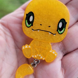 May include: A yellow, sparkly, cartoon-style figurine of a Pokemon character with green eyes and a white crescent moon tail. The figurine is attached to a silver chain.