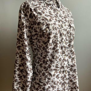 Puede incluir: Una camisa blanca de manga larga con botones y estampado paisley marrón.