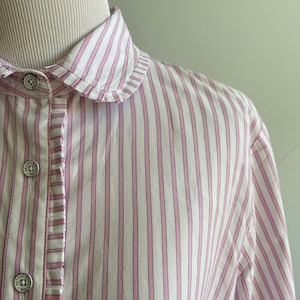 Może przedstawiać: Zbliżenie białej koszuli w różowe paski. Koszula ma falbankowy kołnierz i ozdobne guziki. Pionowe paski są równomiernie rozmieszczone, a guziki mają okrągły wzór.