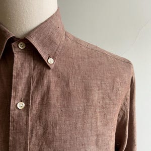 May include: A close-up of a button-down shirt in a reddish-brown color. The shirt has a button-down collar and white buttons. The fabric has a textured appearance.