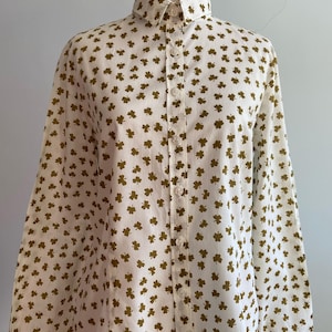 Op de afbeelding: Een witte button-down shirt met een herhalend patroon van kleine, olijfgroene klavers. Het shirt met lange mouwen wordt getoond op een mannequin, met de kraag en de voorkant met knopen.