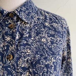 Peut inclure: Gros plan sur une chemise bleu marine &agrave; boutons avec un motif floral cr&egrave;me. La chemise a une poche poitrine et des boutons marron. Le col est visible, et le tissu semble &ecirc;tre du coton.