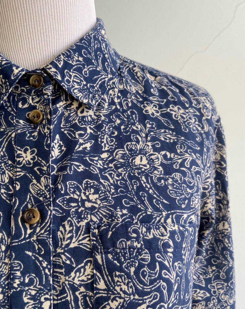 Peut inclure: Gros plan sur une chemise bleu marine &agrave; boutons avec un motif floral cr&egrave;me. La chemise a une poche poitrine et des boutons marron. Le col est visible, et le tissu semble &ecirc;tre du coton.