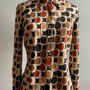 May include: A long-sleeved button-down shirt featuring a repeating geometric pattern of overlapping circles in shades of dark brown, tan, cream, and burnt orange. The shirt has a standard collar and a front placket with visible buttons.