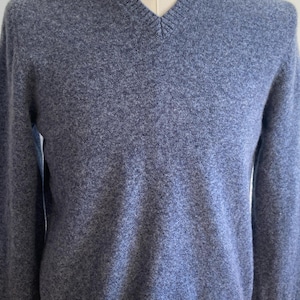 May include: A blue V-neck sweater with a ribbed hem and cuffs. The sweater is made of a soft, textured material and is displayed on a mannequin. The overall style is classic and suitable for various occasions.