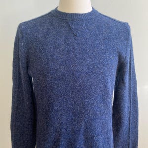 Puede incluir: Un suéter azul de cuello redondo con un tejido texturizado. El suéter de manga larga se muestra en un maniquí. El escote presenta un pequeño detalle triangular. El suéter es de color sólido.