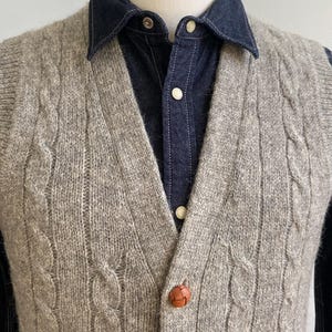 May include: A light gray cable-knit sweater vest with brown leather buttons. The vest is worn over a dark blue denim shirt with white buttons. The overall style is classic and preppy.