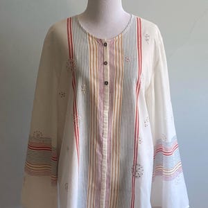 Puede incluir: Blusa de manga larga color crema con rayas verticales rojas, azules, amarillas y marrones. La blusa tiene cuello redondo, cierre de botones en la parte delantera y detalles florales bordados. Las mangas tienen rayas horizontales en los puños.