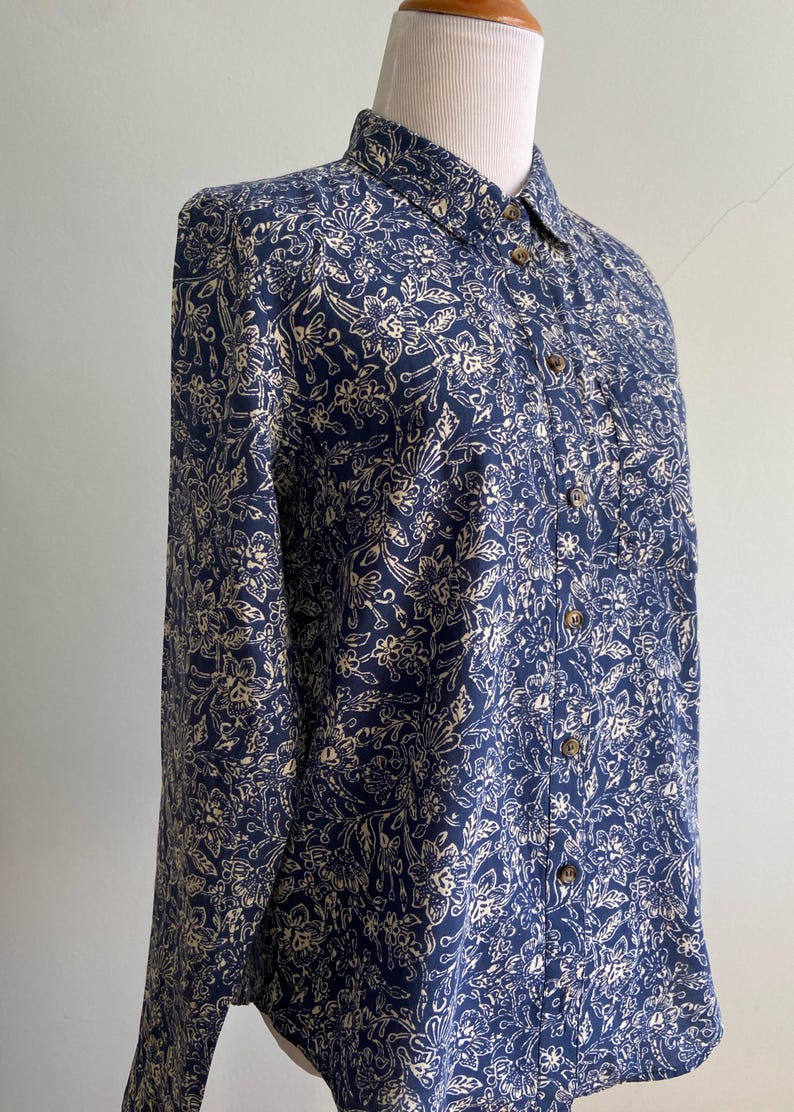 Peut inclure: Chemise &agrave; manches longues bleu marine avec un motif floral cr&egrave;me. La chemise a un col classique et des boutons en bois. Le tissu semble l&eacute;ger, id&eacute;al pour les temp&eacute;ratures plus chaudes. La chemise est pr&eacute;sent&eacute;e sur un mannequin.
