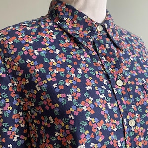 Könnte beinhalten: Ein marineblaues Hemd mit durchgehender Knopfleiste und einem dichten, mehrfarbigen Blumenmuster aus kleinen roten, gelben, rosafarbenen und weißen Blüten sowie grünen Blättern. Das Hemd verfügt über einen klassischen Kragen und weiße Knöpfe.