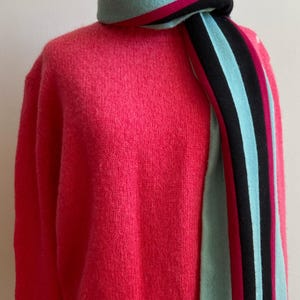 May include: A bright pink sweater with a matching scarf. The scarf has stripes of teal, black, and pink. The sweater and scarf are displayed on a mannequin.