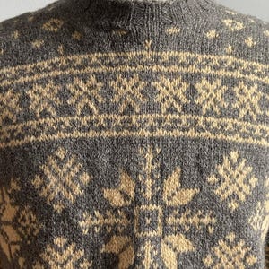 May include: A charcoal gray knit sweater featuring a cream-colored fair isle pattern with repeating snowflake and geometric motifs. The garment has a ribbed mock neck collar and a textured wool-like appearance with long sleeves and a fitted cut.