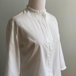 以下が含まれることがあります： 白い長袖ブラウス。フリル襟とボタン留めが特徴です。ブラウスは体にフィットしたボディと、少し広がった裾を持っています。