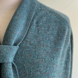 May include: A teal-colored knit sweater with a shawl collar and a tie detail. The sweater has a textured appearance with flecks of brown and blue throughout the fabric. The close-up shot highlights the soft, cozy material and the garment's design.