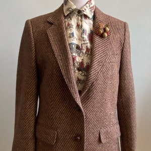 Puede incluir: Una chaqueta de tweed marrón con un solo botón de cierre. La chaqueta lleva una camisa estampada debajo con un diseño floral y un pequeño alfiler de bellota en la solapa.