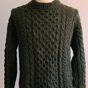 Puede incluir: Jersey de punto verde oscuro con un patrón texturizado y cuello redondo. El jersey tiene un diseño de punto trenzado en las mangas y los lados, y un patrón de panal en la parte delantera. El hilo tiene motas de color.