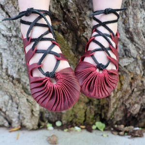 May include: A pair of burgundy leather sandals with black crisscross laces. The sandals have a unique pleated design at the toe and ankle straps. The shoes are designed for comfort and style.