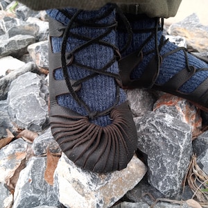 May include: Close-up of a pair of black leather shoes with a unique design. The shoes have a lace-up closure and are worn with blue socks. The shoes are standing on a bed of gray rocks.