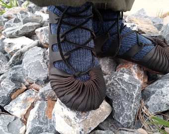 May include: Close-up of a pair of black leather shoes with a unique design. The shoes have a lace-up closure and are worn with blue socks. The shoes are standing on a bed of gray rocks.