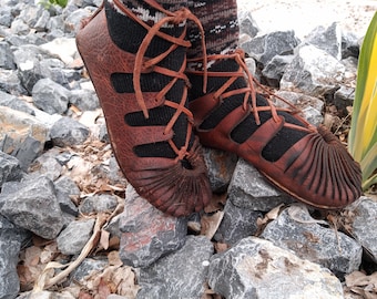 May include: Close-up of a pair of brown leather boots with black laces and a unique design. The boots are worn on a rocky surface.