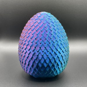Puede incluir: Un huevo de dragón azul y morado con una superficie texturizada y escamosa. El huevo está hecho de un material impreso en 3D.