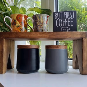 May include: A dark-stained wooden riser shelf holding two ceramic mugs with floral patterns and a black sign with white text that reads BUT FIRST COFFEE. Below the shelf are two matte canisters with wooden lids, one black and one dark grey.