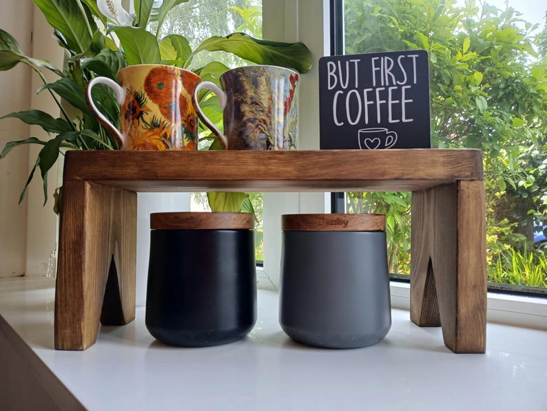 May include: A dark-stained wooden riser shelf holding two ceramic mugs with floral patterns and a black sign with white text that reads BUT FIRST COFFEE. Below the shelf are two matte canisters with wooden lids, one black and one dark grey.