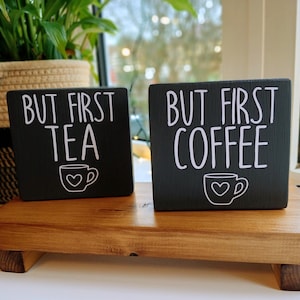 May include: Two rectangular black wooden signs with white text and a small cup graphic. The smaller sign reads "BUT FIRST TEA" and the larger sign reads "BUT FIRST COFFEE". Each cup graphic features a small heart in the centre of the cup.