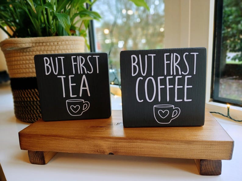 May include: Two rectangular black wooden signs with white text and a small cup graphic. The smaller sign reads "BUT FIRST TEA" and the larger sign reads "BUT FIRST COFFEE". Each cup graphic features a small heart in the centre of the cup.