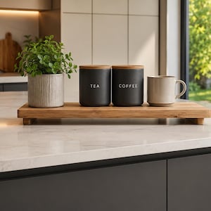 May include: A wooden serving tray displays a small potted plant, two black canisters marked "TEA" and "COFFEE", and a beige mug. The canisters have wooden lids. The items are arranged on a white countertop in a kitchen.
