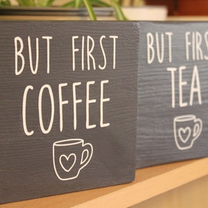 May include: Two square dark grey wooden signs with white lettering and graphics. One sign reads BUT FIRST COFFEE with a mug icon featuring a heart. The second sign reads BUT FIRST TEA with a matching mug icon featuring a heart. Both signs have a visible wood grain texture.