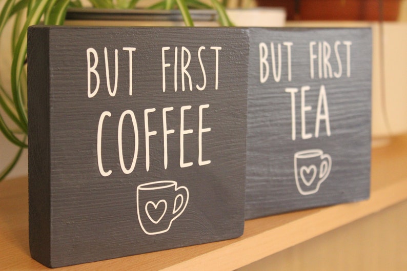 May include: Two square dark grey wooden signs with white lettering and graphics. One sign reads BUT FIRST COFFEE with a mug icon featuring a heart. The second sign reads BUT FIRST TEA with a matching mug icon featuring a heart. Both signs have a visible wood grain texture.