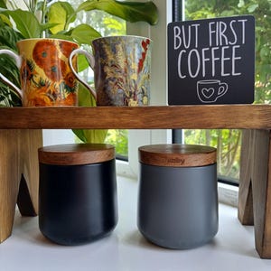 May include: A wooden riser shelf holding two ceramic mugs with floral patterns and a black sign with white text reading BUT FIRST COFFEE and a heart-shaped cup icon. Below the shelf are two matte canisters, one black and one grey, with wooden lids.