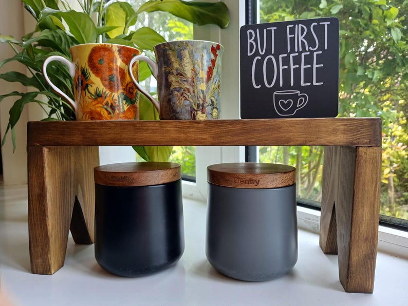 May include: A wooden riser shelf holding two ceramic mugs with floral patterns and a black sign with white text reading BUT FIRST COFFEE and a heart-shaped cup icon. Below the shelf are two matte canisters, one black and one grey, with wooden lids.