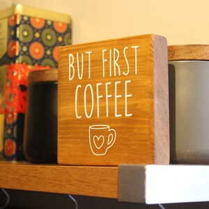 May include: A square wooden sign with a medium brown finish featuring the white text BUT FIRST COFFEE and a line drawing of a coffee mug with a heart in the centre. The sign is positioned upright on a shelf between two cylindrical containers.