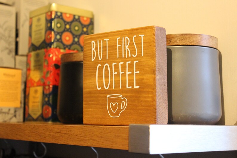 May include: A square wooden sign with a medium brown finish featuring the white text BUT FIRST COFFEE and a line drawing of a coffee mug with a heart in the centre. The sign is positioned upright on a shelf between two cylindrical containers.