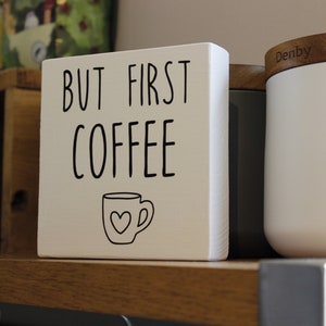 May include: A square white wooden block sign featuring the black text "BUT FIRST COFFEE" in a simple sans-serif font. Below the text is a black line drawing of a coffee mug with a small heart shape centred on the front of the mug.