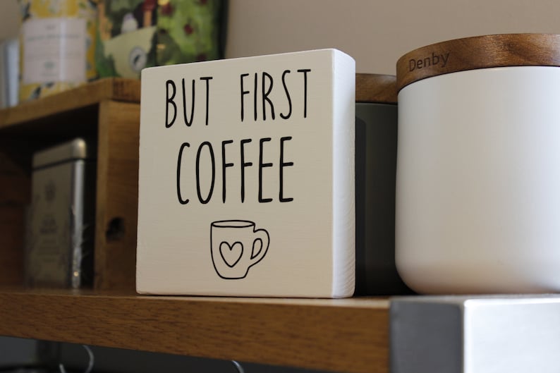 May include: A square white wooden block sign featuring the black text "BUT FIRST COFFEE" in a simple sans-serif font. Below the text is a black line drawing of a coffee mug with a small heart shape centred on the front of the mug.